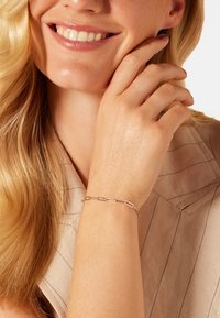 Zilveren armband met verlengde schakels, gedragen op een hand tegen een lichtbruine stoffen achtergrond. Gladde textuur, minimalistisch ontwerp.