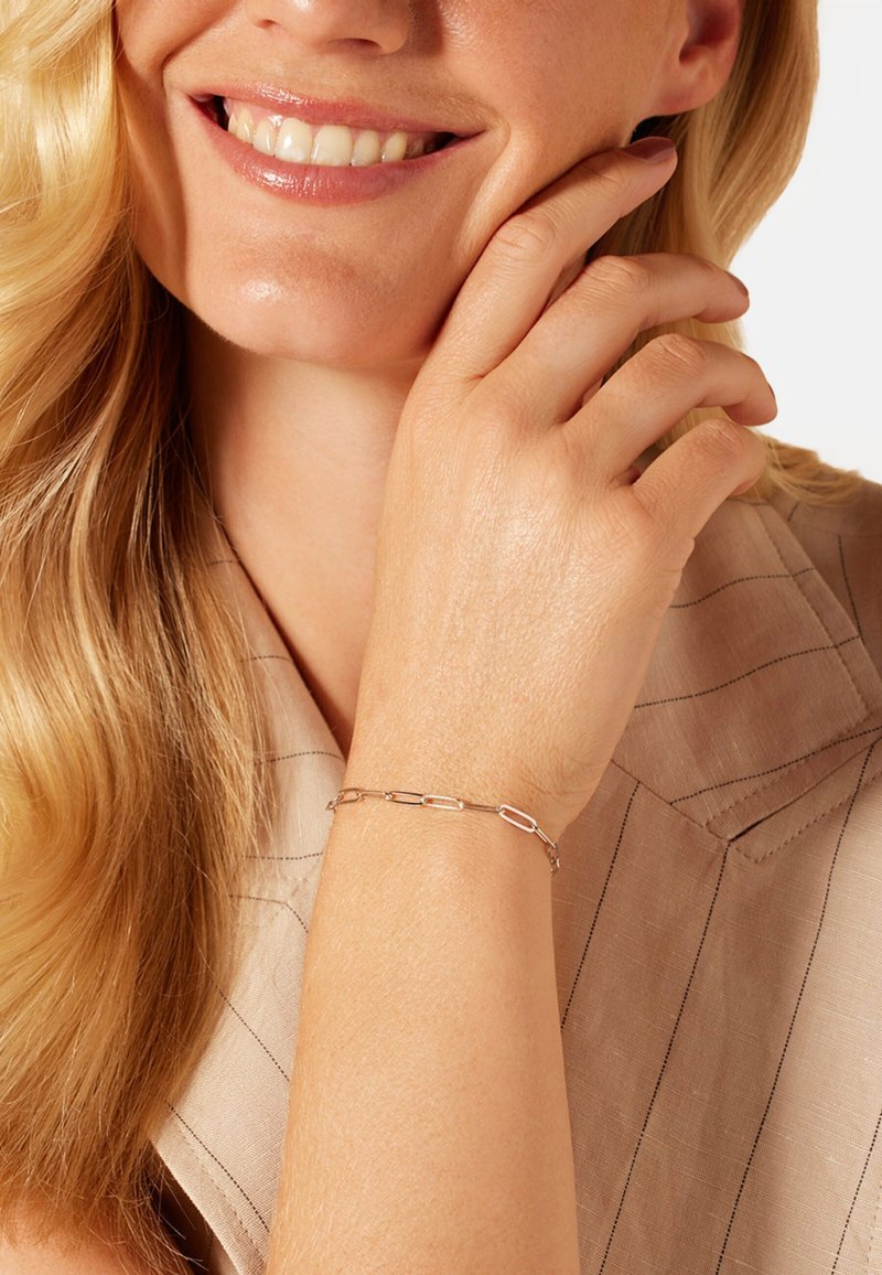Zilveren armband met verlengde schakels, gedragen op een hand tegen een lichtbruine stoffen achtergrond. Gladde textuur, minimalistisch ontwerp.