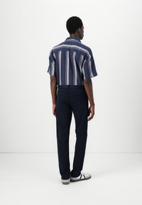 Cămașă cu mânecă scurtă, în dungi, de culoare bleumarin cu accente albe și roșii, asortată cu pantaloni închiși și adidași. Textura pare netedă și ușoară.