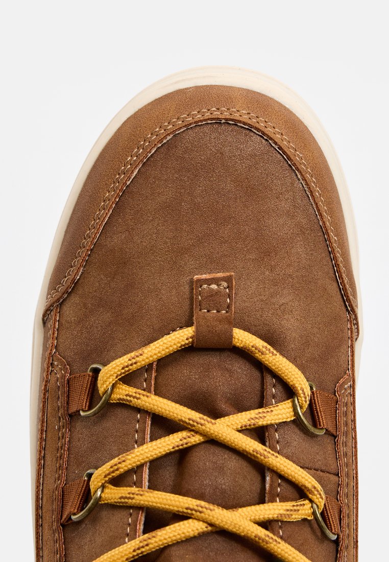 Braune Lederstiefel mit gelben Schnürsenkeln, abgerundeter Zehenpartie und cremefarbener Sohle. Mit Metallösen und genähten Details entlang der Kanten.