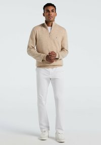 Beige Reißverschluss-Pullover aus weichem Strickstoff, kombiniert mit weißen geraden Hosen und weißen Turnschuhen, steht vor einem neutralen Hintergrund.