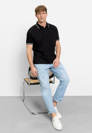 Man zit op een metalen stoel met een gevlochten zitting, draagt een zwart poloshirt, lichtblauwe spijkerbroek en witte sneakers tegen een effen witte achtergrond.