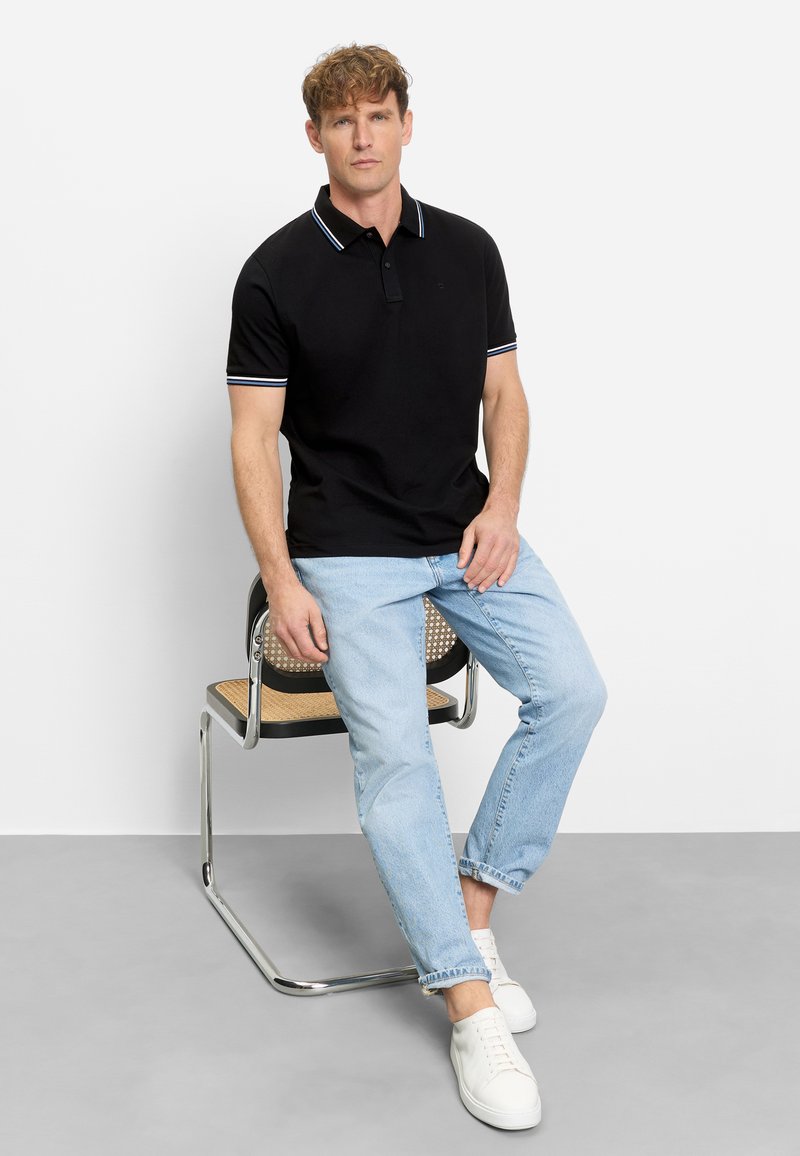 Man zit op een metalen stoel met een gevlochten zitting, draagt een zwart poloshirt, lichtblauwe spijkerbroek en witte sneakers tegen een effen witte achtergrond.