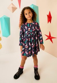 Marineblauwe jurk met lange mouwen en een feestelijke print met eenhoorns, bomen en sneeuwmanneken. Combinatie met zwarte laarzen en roze gebreide sokken.