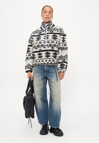Fleece mikina s geometrickým vzorem v námořnické modři a bílé barvě, čtvrtový zip vpřed, vysoký límec a jednoduchý modrý zip. Dvojice světle modrých džínů a černá taška.