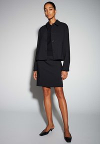 Zwarte cropped jas met knopen, gecombineerd met een aansluitende zwarte rok en zwarte pumps met puntige neus, allemaal gemaakt van een gladde stof. Eenvoudig ontwerp.