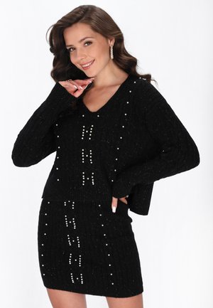 Kvinde smilende iført et sort strikket sæt med sweater og nederdel dekoreret med perleaccents og lodrette mønstre, poserer med én hånd nær hendes hage.