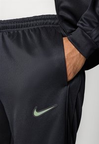 Črne atletske hlače z gladkim materialom, z stransko žepom in vidnim zelenim Nike swoosh logotipom na stegnu.