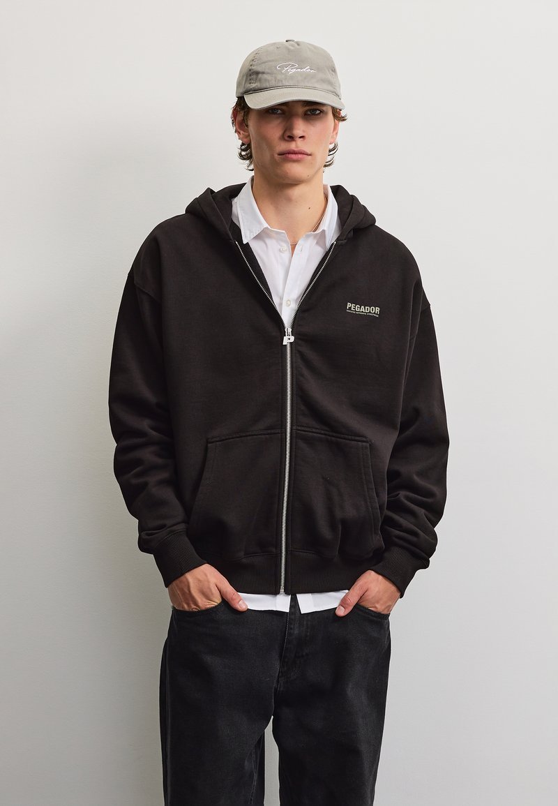 Jonge man met een grijze pet, zwarte zip-up hoodie met voorzakken, wit shirt en donkere broek, staand tegen een effen lichte achtergrond.