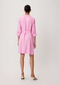 Abito rosa alla ginocchia con vita regolabile, maniche lunghe arrotolate ai polsini e una texture liscia. Presenta una fessura sul retro.