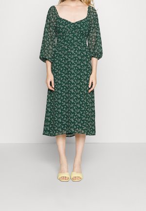 Zielona sukienka midi w kwiaty z kwadratowym dekoltem, przezroczystymi bufkami na rękawach i zwiewną sylwetką. W połączeniu z żółtymi klapkami na płaskim obcasie.