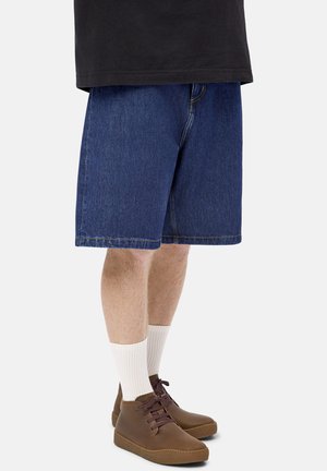 Man wearing blue denim shorts, white crew socks, and brown leather lace-up casual shoes standing against a plain background.