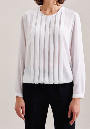 Vrouw draagt een wit blouse met lange mouwen met verticale plooien aan de voorkant en zwarte broek, staand tegen een effen achtergrond.