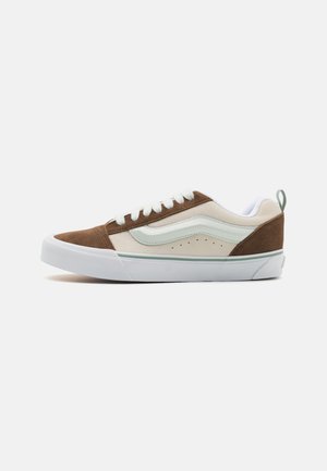 Zapatilla baja de ante marrón y textil crema con suela de goma blanca, que presenta un acento en verde menta y un detalle de logo clásico en el lateral.