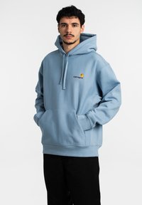 Sudadera de algodón azul con un bolsillo frontal, capucha con cordón y logotipo bordado en amarillo y burdeos. Corte holgado con puños y dobladillo acanalados.