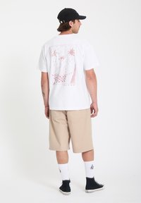 Homme portant un t-shirt graphique blanc, un short beige, une casquette noire, des chaussettes blanches mi-mollet et des baskets montantes noires, debout devant un fond uni.