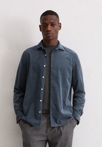 Ein Mann steht mit den Händen in den Taschen, trägt ein dunkelgraues Hemd über einem grauen Pullover und graue Hosen vor einer schlichten weißen Wand.