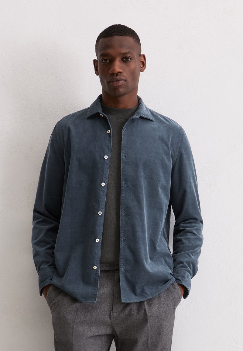 Ein Mann steht mit den Händen in den Taschen, trägt ein dunkelgraues Hemd über einem grauen Pullover und graue Hosen vor einer schlichten weißen Wand.