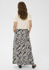 Kvinde med bølget brunt hår iført en lys cremefarvet kortærmet bluse og en lang sort, beige og hvid zebra-print nederdel sammen med sorte flade sko.