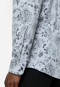 Langärmliges Hemd aus hellgrauem Stoff mit schwarzem Paisleymuster. Mit schwarzen Knöpfen an den Manschetten und glatter, flacher Textur.