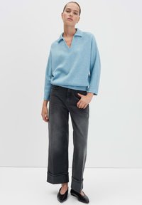 Vrouw in een lichtblauwe lange mouw trui, donkere wijde broek met omslag en zwarte puntige ballerina's, staande tegen een effen witte achtergrond.