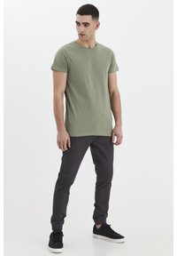Maslinasto zelena pamučna majica s okruglim izrezom, u kombinaciji s tamnosivim trenirkama s elastičnim manžetama. Crne tenisice upotpunjuju ovaj outfit.