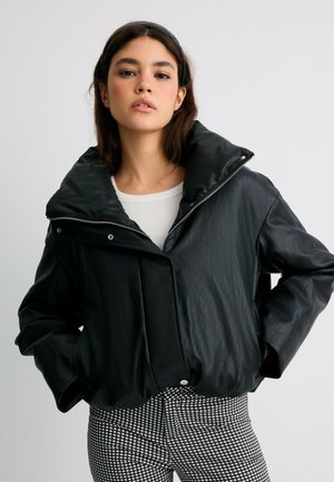 Jeune femme portant une veste en cuir noire à large col, un top blanc et un pantalon à carreaux noir et blanc, debout les mains dans les poches.