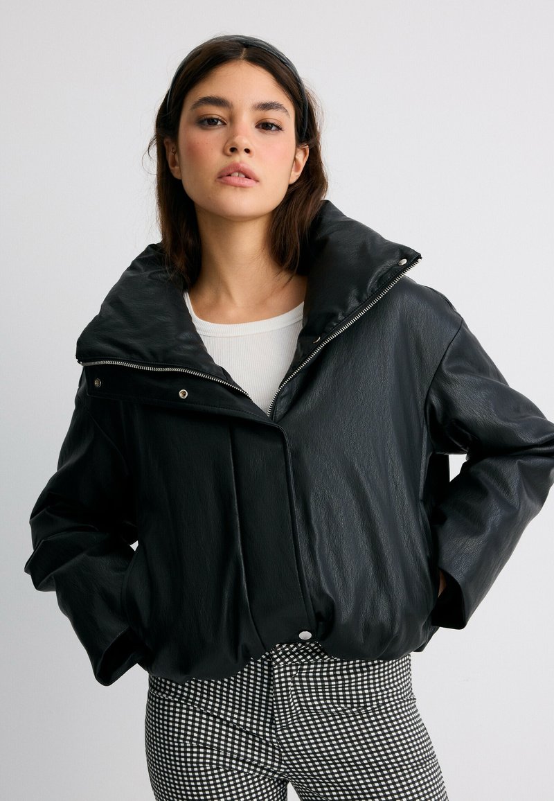 Jeune femme portant une veste en cuir noire à large col, un top blanc et un pantalon à carreaux noir et blanc, debout les mains dans les poches.