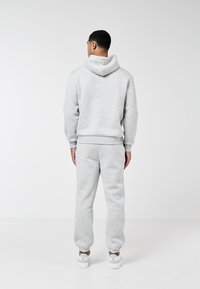 Bărbat care poartă hanorac gri deschis și pantaloni de trening, stând cu spatele la fundal alb simplu.