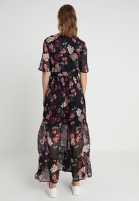 Robe maxi noire à fleurs avec manches courtes, volants en étages et taille cintrée ; décorée de fleurs roses et blanches sur un tissu transparent.