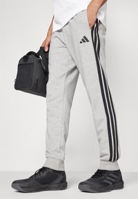 Grå joggebukser med sorte striper, med et Adidas-logo. Stoffet ser mykt ut, med ribbestrikkede mansjetter og lommer. En svart veske holdes i hånden.