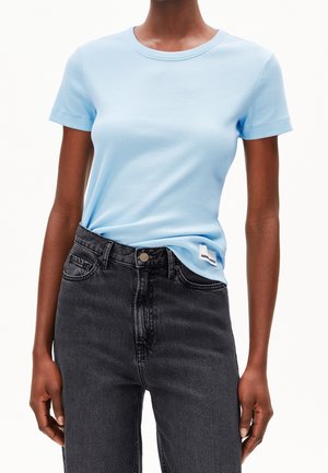Personne portant une chemise bleu clair à manches courtes rentrée dans un jean noir taille haute, les mains relâchées le long du corps.