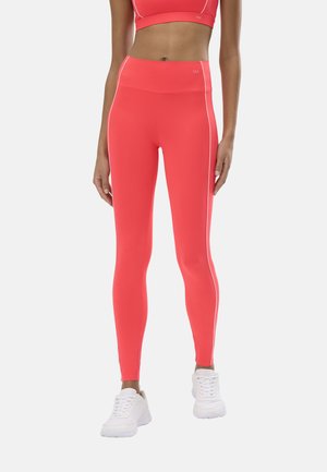 Mujer que lleva leggings de cintura alta color coral brillante con franjas blancas laterales y un sujetador deportivo a juego, combinados con zapatillas deportivas blancas.