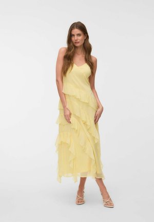 Femme aux longs cheveux bruns portant une robe jaune sans manches à larges couches et des talons beiges à brides, debout devant un fond blanc uni.