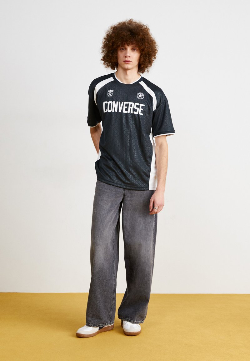 Jersey negro de manga corta con detalles en blanco y la marca "Converse", combinado con jeans de pierna ancha grises y zapatillas blancas. Fondo plano.