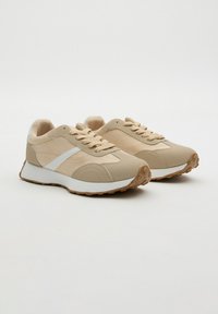 Zapatillas deportivas beige con franja lateral blanca, cierre de cordones y suelas de goma marrón texturizada sobre un fondo liso.