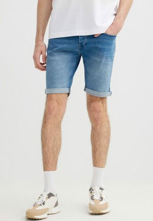 Hombre con pantalones cortos vaqueros azules remangados, calcetines blancos hasta la mitad de la pierna, zapatillas beige y blancas, de pie con una mano en el bolsillo.