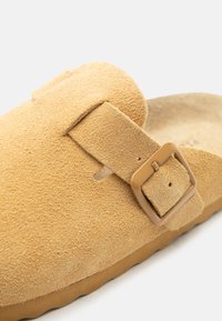 Sabot en daim marron clair avec large bride et boucle rectangulaire, avec semelle texturée pour adhérence et confort.