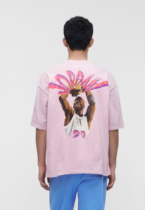 Person, die ein hellrosa T-Shirt mit einer Grafik eines Basketballspielers, der wirft, und dem Wort „Jordan“ in leuchtendem, stilisiertem Schriftzug auf dem Rücken trägt.