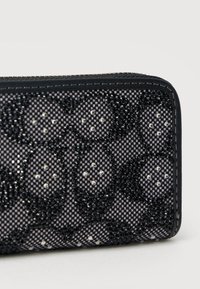 Κοντινό πλάνο ενός πορτοφολιού με φερμουάρ, φτιαγμένο από καρό ύφασμα σε μαύρο και γκρι χρώμα, διακοσμημένο με ανθικά σχέδια από μαύρες και ασημένιες χάντρες και μαύρα ραμμένα άκρα.