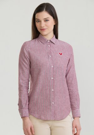 Femme portant une chemise à boutons rayée verticalement rouge et blanche avec un petit emblème de cœur rouge sur la poitrine gauche, se tenant devant un fond uni.