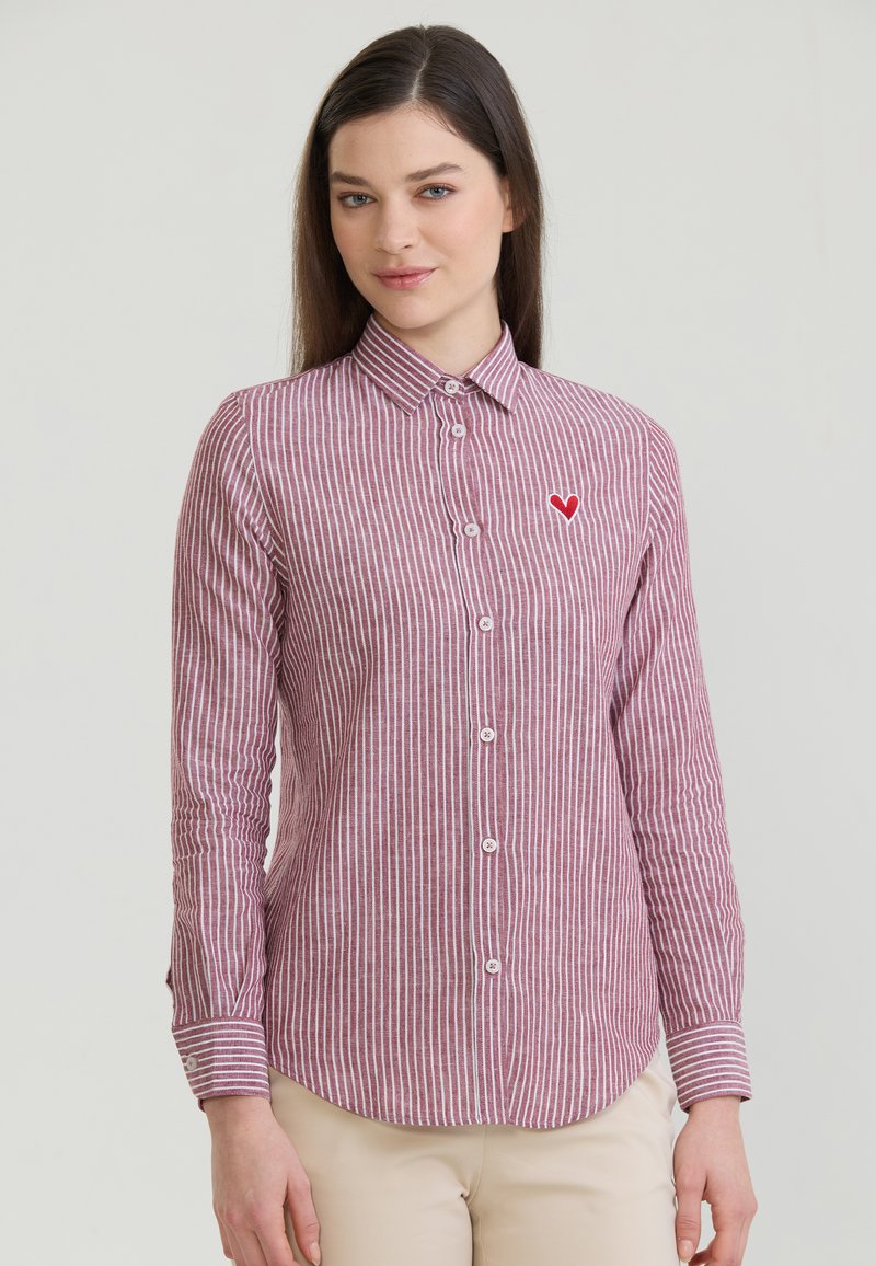 Femme portant une chemise à boutons rayée verticalement rouge et blanche avec un petit emblème de cœur rouge sur la poitrine gauche, se tenant devant un fond uni.