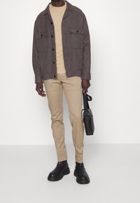 Szara bawełniana kurtka z zapięciem na guziki, beżowy t-shirt, beżowe spodnie dopasowane, czarne botki do kostki. Trzyma małą czarną torebkę.