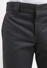 Pantalon noir à texture lisse, doté d'un devant plat et de passants pour ceinture, conçu pour une coupe ajustée. Détails de couture minimaux.