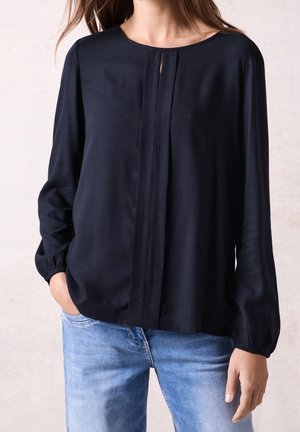 Mujer con una blusa negra de manga larga con un detalle de pliegue vertical sobre unos vaqueros azul claro, de pie con una mano en el bolsillo.