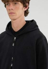 Felpa nera con zip frontale, realizzata in tessuto morbido. Presenta un cappuccio e un logo in rilievo sul petto. Design aderente.