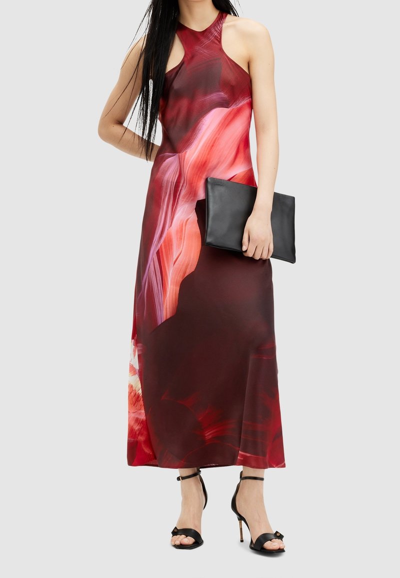 Femme portant une robe longue sans manches avec des motifs abstraits rouges et roses, tenant une pochette noire et portant des sandales à talons hauts noires.