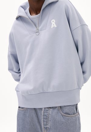 Personne portant un sweat-shirt violet clair à fermeture quart de zip avec un logo blanc et un jean en denim bleu clair, debout devant un fond uni.