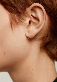 Pendiente de aro dorado con un pequeño detalle brillante, llevado en el lóbulo de una persona con cabello corto de color castaño rojizo, destacando la oreja.