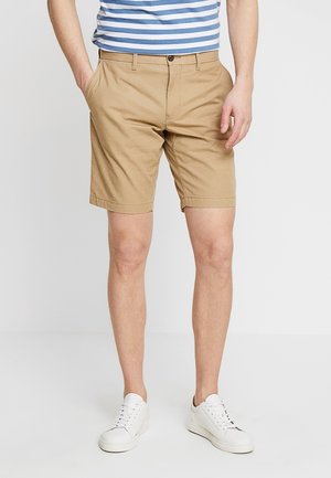 Mann trägt beige knielange Shorts, weiße Sneaker und ein blau-weiß gestreiftes Hemd, steht mit den Händen in den Taschen.